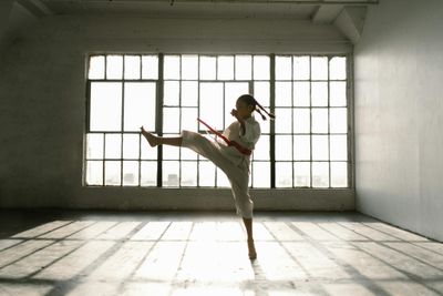 A young girl in a martial arts uniform practicing a high kick in a sunlit dojo.