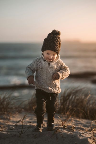 Adorable toddler in warm knitwear exploring a sandy beach with a joyful smile at sunset.