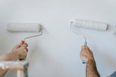 Close-up of two individuals painting a wall with paint rollers indoors.