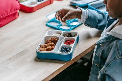 From above side view of crop unrecognizable ethnic schoolkid at table with lunch container full of tasty food
