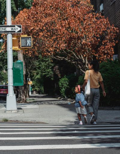 Full body back view of Asian anonymous mother with bag and little girl holding hands while walking on crosswalk on street