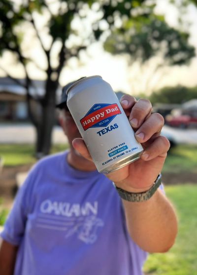 A person holding a can of Happy Dad Texas Seltzer with the Fruit Punch flavor prominently displayed on the can's label. The b