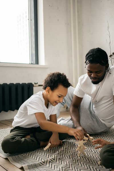 Pensive African American father in casual clothes sitting on floor and playing in board game with little son at home in dayti