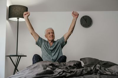 Senior man in pajamas waking up with arms stretched in a cozy bedroom setting.