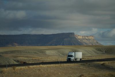 Truck driving through vast desert landscape with dramatic cloudy sky and mountain backdrop.