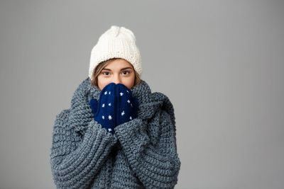 Young beautiful fair-haired woman in knited hat sweater and mittens smiling on grey.