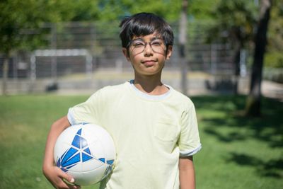 Young boy with glasses holding a soccer ball in a sunny park in Portugal.