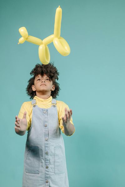 Young girl in overalls playing with a yellow balloon animal against a blue background.