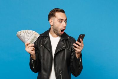 Young man in black leather jacket white t-shirt holding fan of cash money in dollar banknotes, cellphone isolated on blue wal