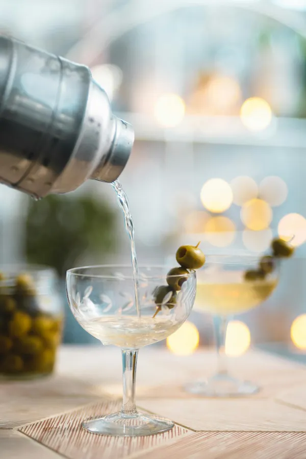 A classic vodka martini being poured from a shaker into a glass garnished with green olives.