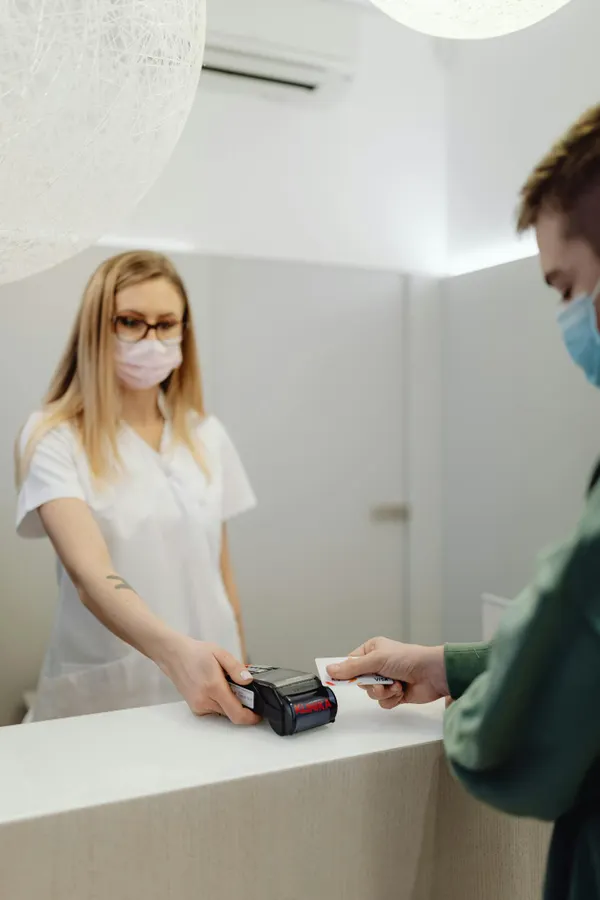 A masked patient pays at a clinic counter, using a card terminal handled by a healthcare professional.