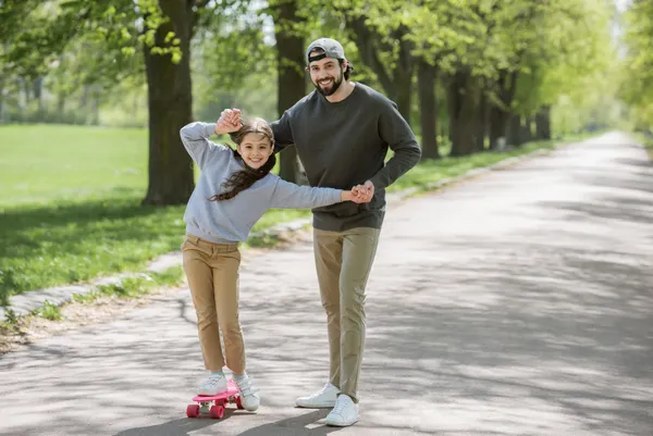 smiling father helping daughter to ride on skateboard in park