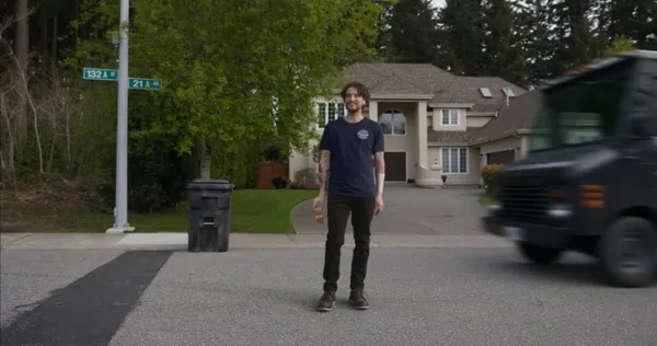A man smiles while standing casually in a suburban street, unaware of an oncoming delivery truck speeding toward him.