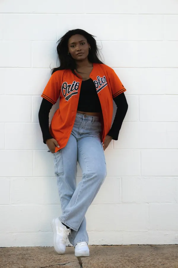 Confident woman in orange sports jersey and jeans posing against a white wall outdoors.