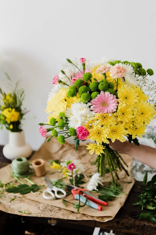Crop unrecognizable florist composing chrysanthemum bouquet in workshop near messy table covered with cut leaves and floristr
