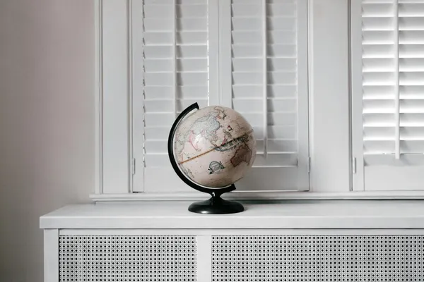 Elegant globe on a minimalist white desk against shuttered windows in modern interior design.