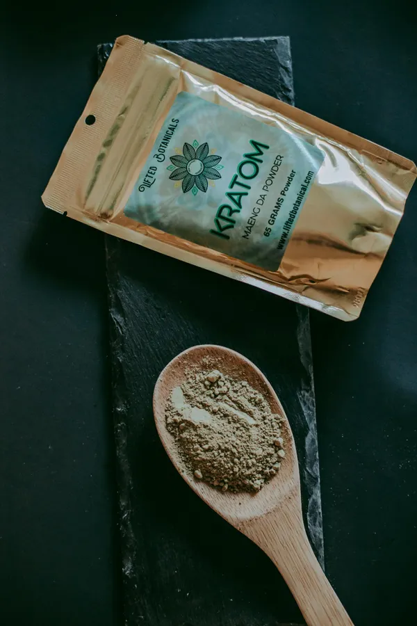 Kratom Maeng Da powder displayed with packet and wooden spoon on a dark surface.