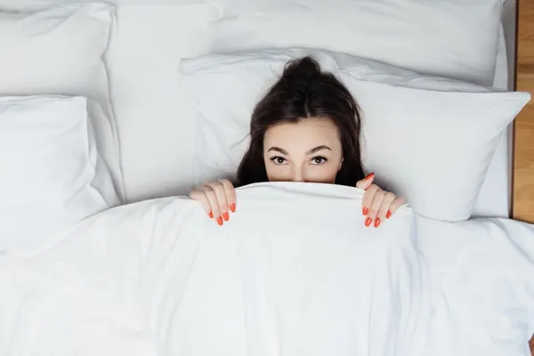 Portrait of surprised lady laying in the bed under white duvet and covers her body