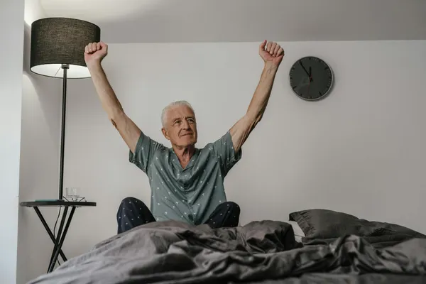 Senior man in pajamas waking up with arms stretched in a cozy bedroom setting.