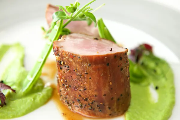 Sliced pork tenderloin with herb garnish and green puree on a white plate.