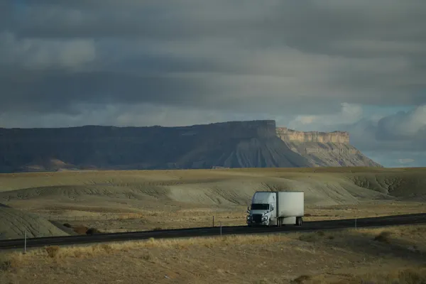 Truck driving through vast desert landscape with dramatic cloudy sky and mountain backdrop.