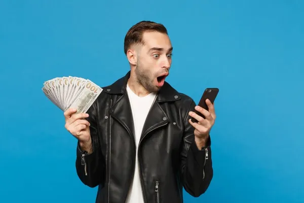 Young man in black leather jacket white t-shirt holding fan of cash money in dollar banknotes, cellphone isolated on blue wal
