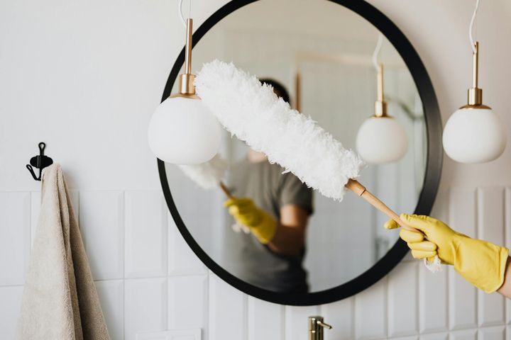 A person cleaning an elegant bathroom mirror with a fluffy duster and yellow gloves.