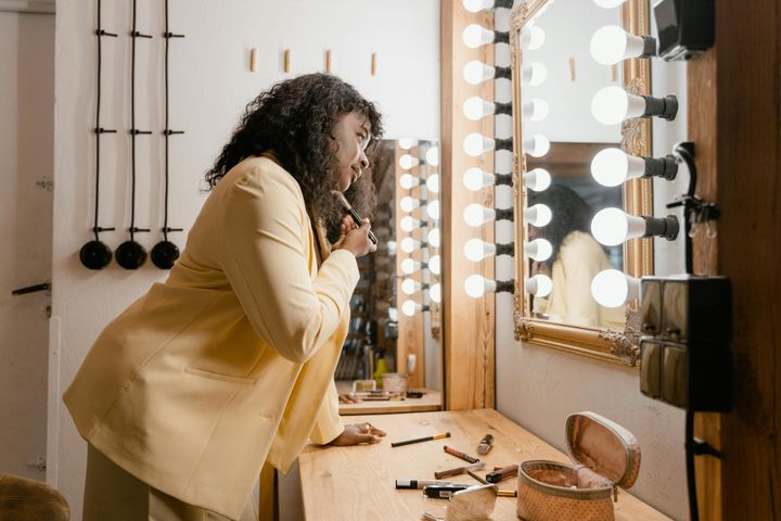African American woman applying makeup at a vanity mirror with lights, wearing a yellow blazer.