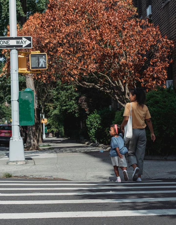 Full body back view of Asian anonymous mother with bag and little girl holding hands while walking on crosswalk on street
