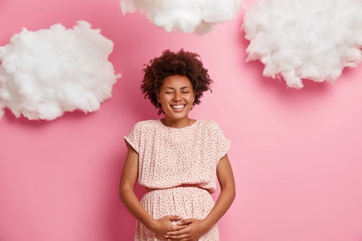 Happy dark skinned pregnant woman touches big belly, cares about future child, smiling expectant mother has good mood, stands