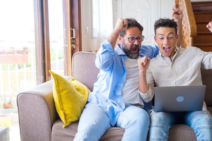 Happy excited grown son and mature 50s father using laptop, celebrating success. two family generations of sport fan watching