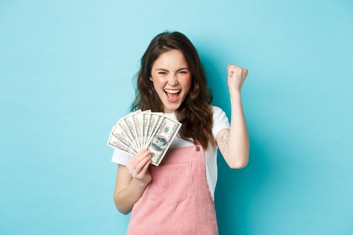 Lucky young woman looks excited, shouting from satisfaction and triumph, winning money, holding dollar bills and making fist