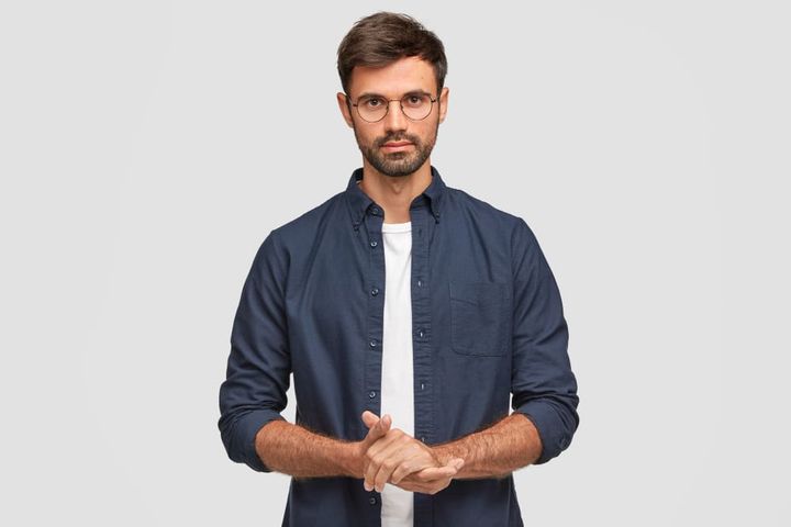 Waist up portrait of handsome serious unshaven male keeps hands together, dressed in dark blue shirt, has talk with interlocu