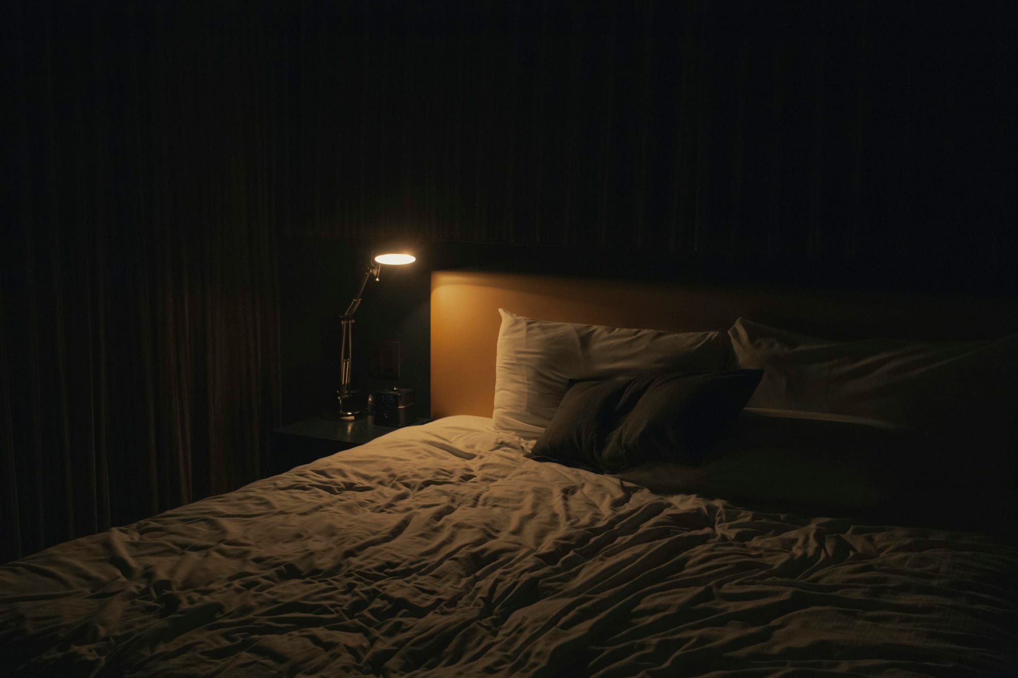 A warm bedside lamp illuminating rumpled ivory bedding in a dark bedroom.