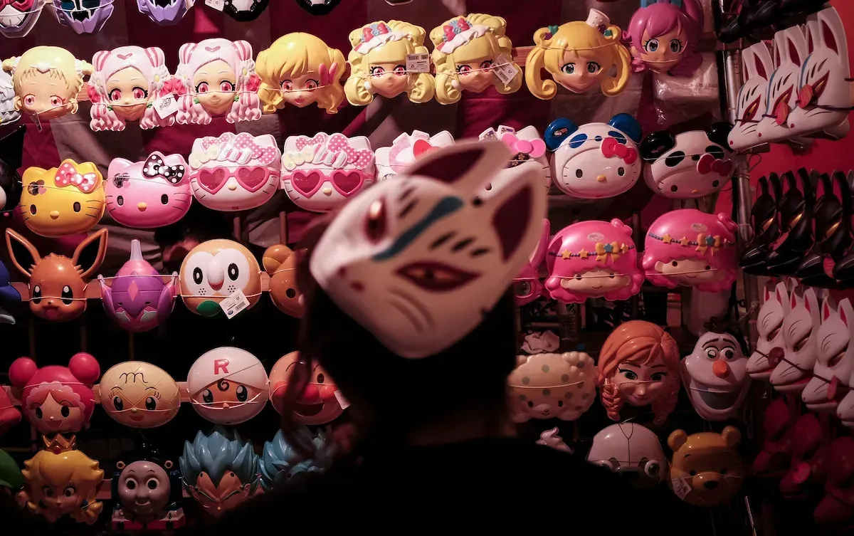 A person wearing a white fox mask in front of shelves of colorful character masks