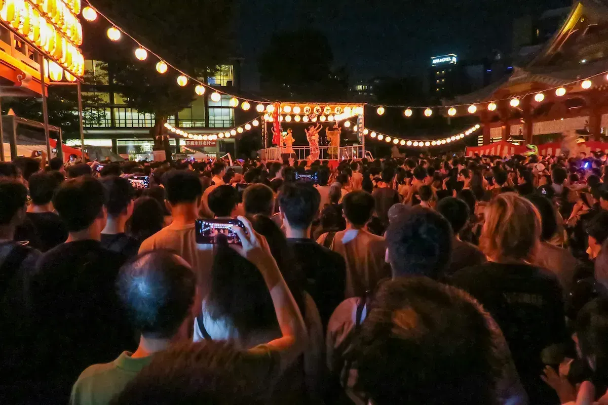 Crowd at a Japanese summer festival at night with lanterns and a stage performance