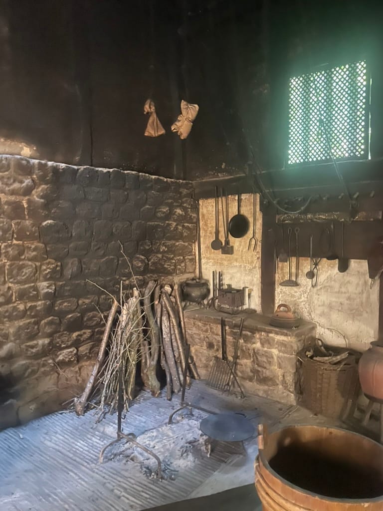 A fifteenth century kitchen at the Weald and Downland museum. A bundle of wood is propped behind the ash of a firepit. Cooking implements hang on the wall.