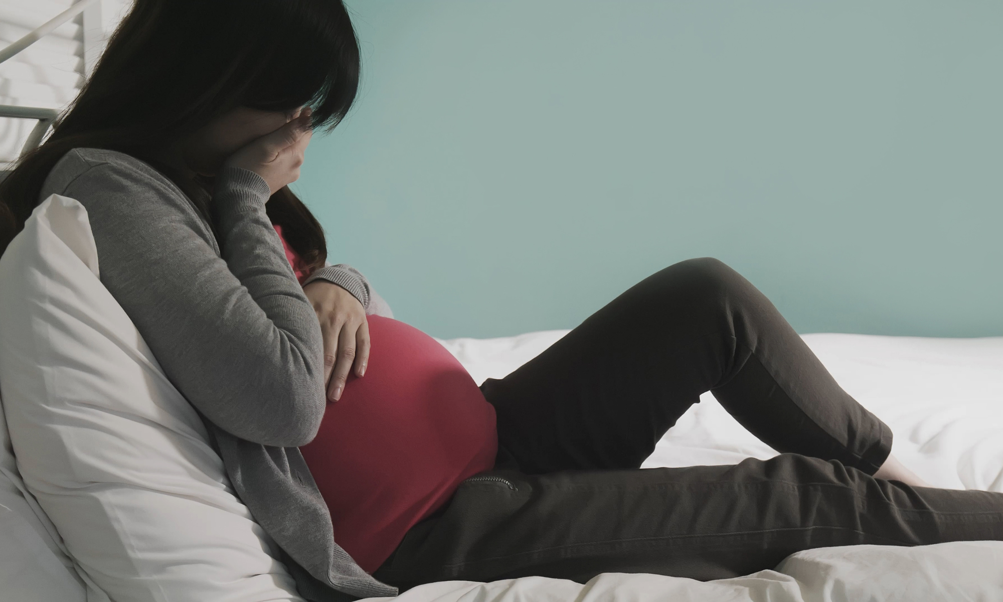 Pregnant woman in red shirt, sad expression, lying in bed.