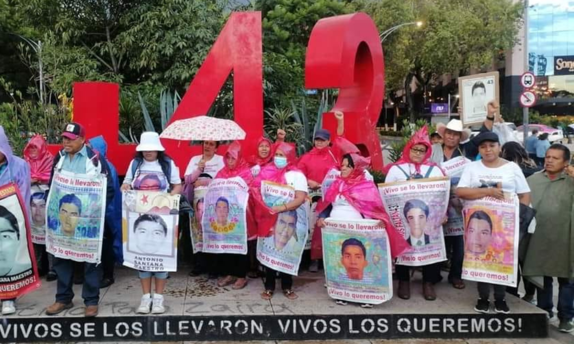 People protesting with signs by a red '43' sculpture, marking the Ayotzinapa students' disappearance.