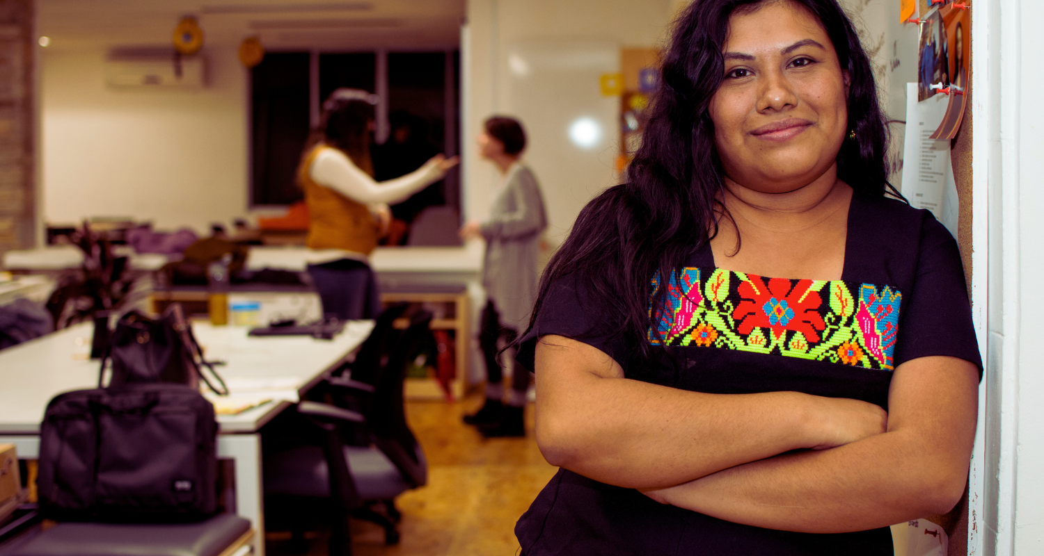 Indigenous Maya woman wearing a traditional embroidered blouse stands with arms crossed in a community workspace, with people collaborating in the background, highlighting leadership, culture, and community organizing.