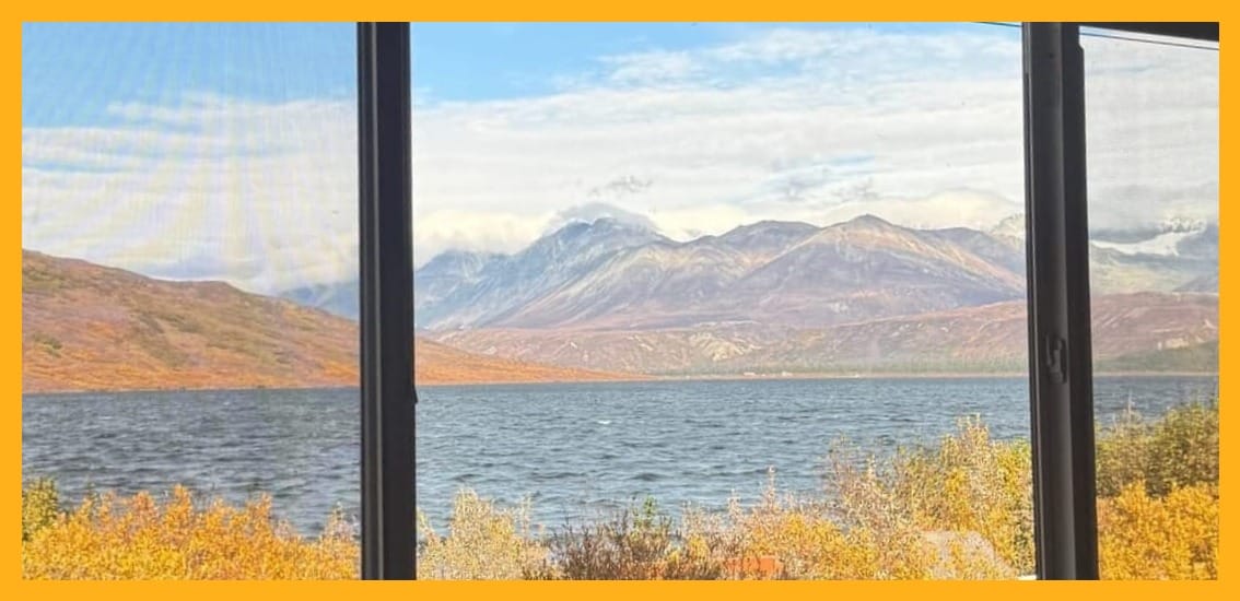 A cabin window overlooking a lake and mountains with autumn colors in Alaska