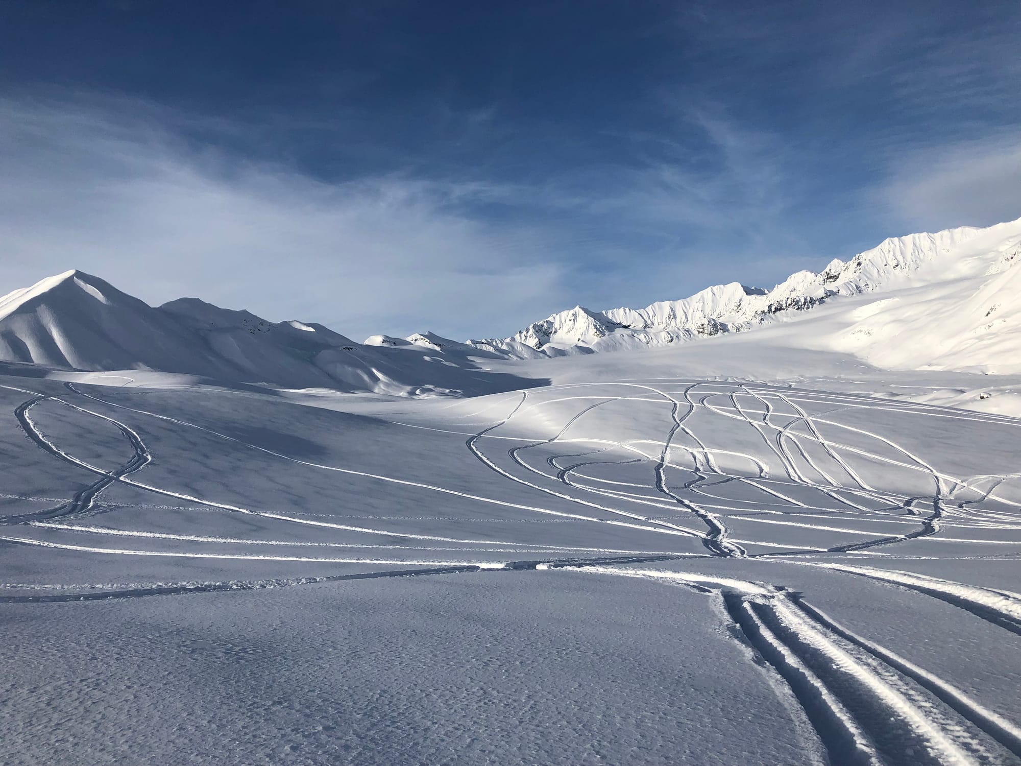 Snow-covered mountain landscape with smooth slopes and multiple snowmachine tracks winding across the terrain under a clear blue sky.
