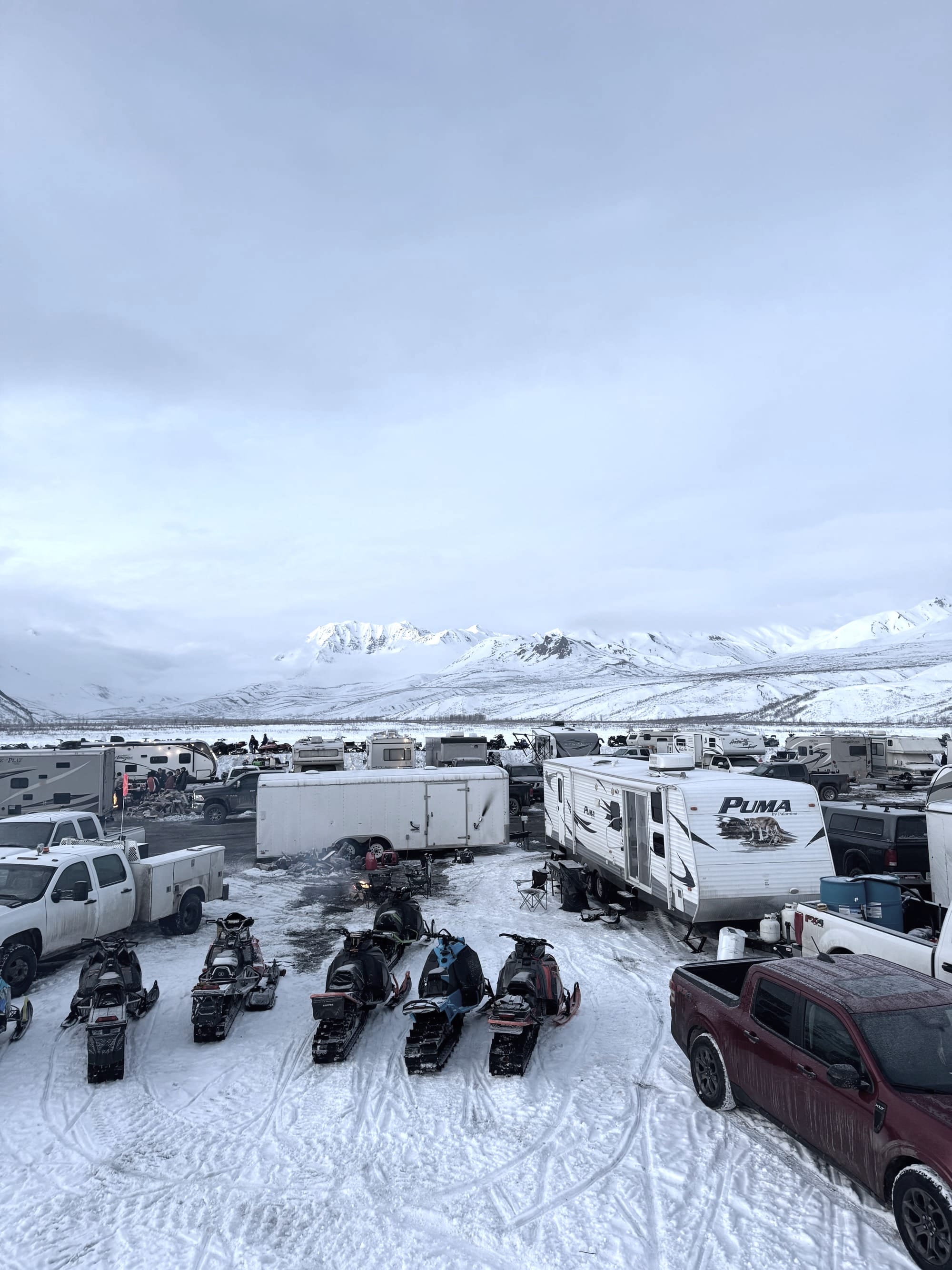Arctic Man Alaska basecamp with snowmachines, trucks and RVs gathered in snowy valley below mountains