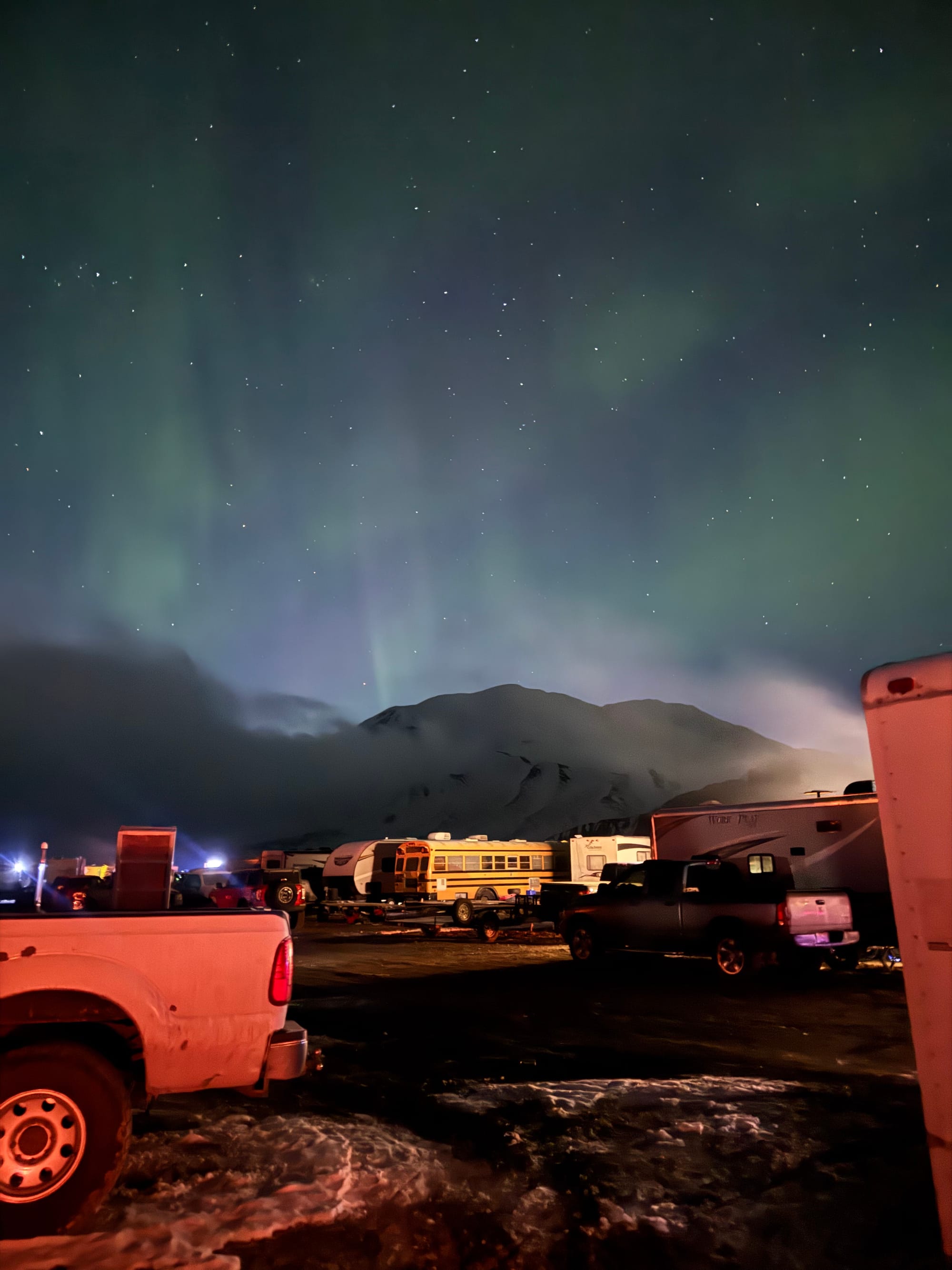 Northern lights glowing in green hues over a snowy mountain at night, with a row of parked trucks, trailers and a bus at Arctic Man under a star-filled sky.