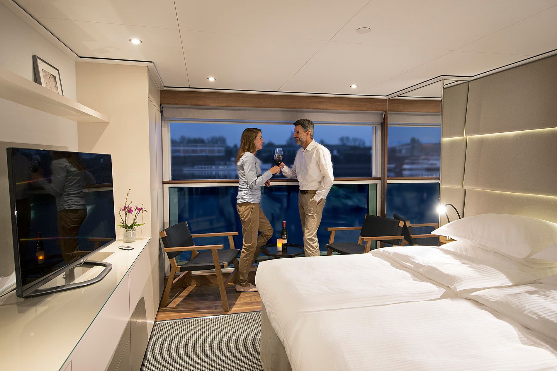 Two white people wearing smart-casual clothing holding glasses of red wine in front of a French balcony
