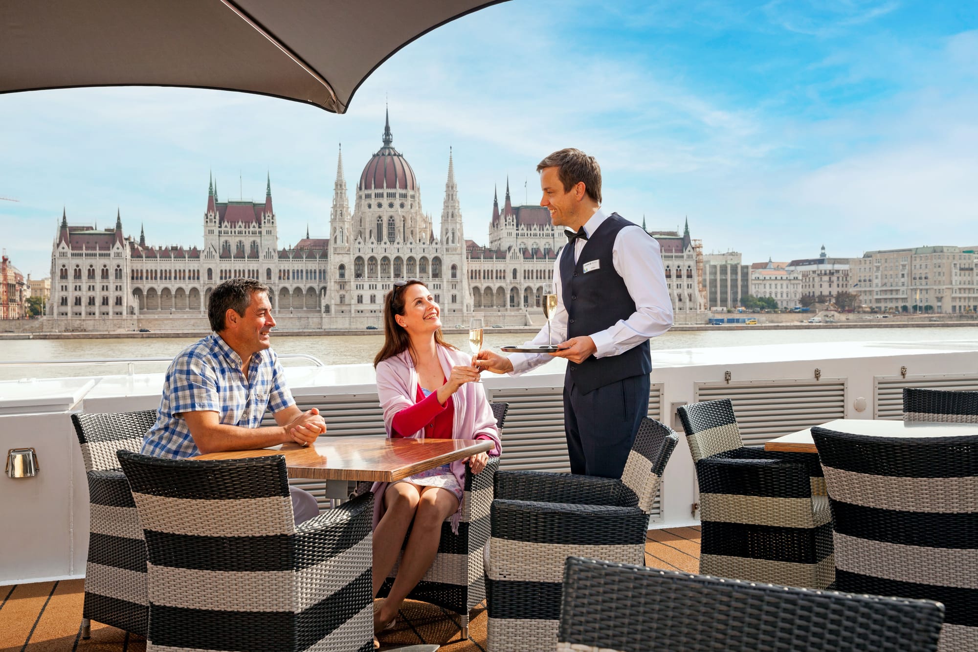 An image of a white couple being served champagne on a river cruise ship, with Budapest in the background