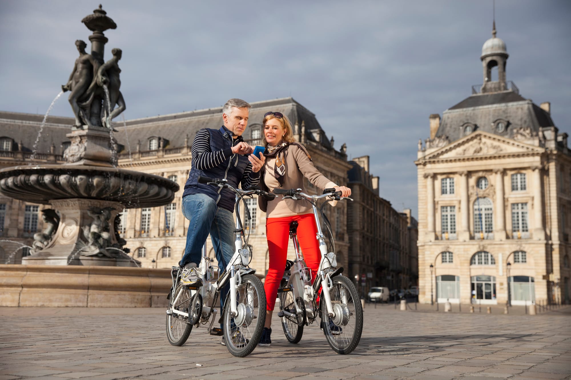 Two white people on e-bikes looking at a phone in a Baroque-style city square