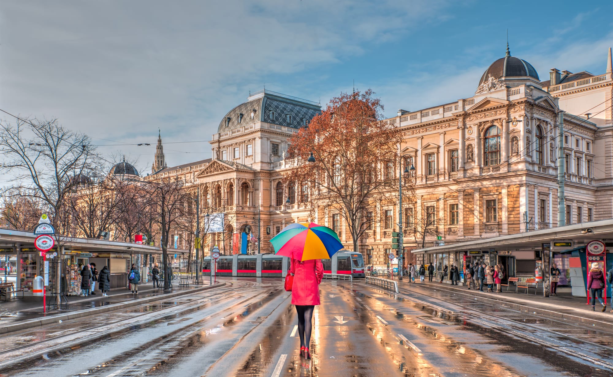 A woman with a colourful umbrella and red coat walking down a road in Vienna after rainfall