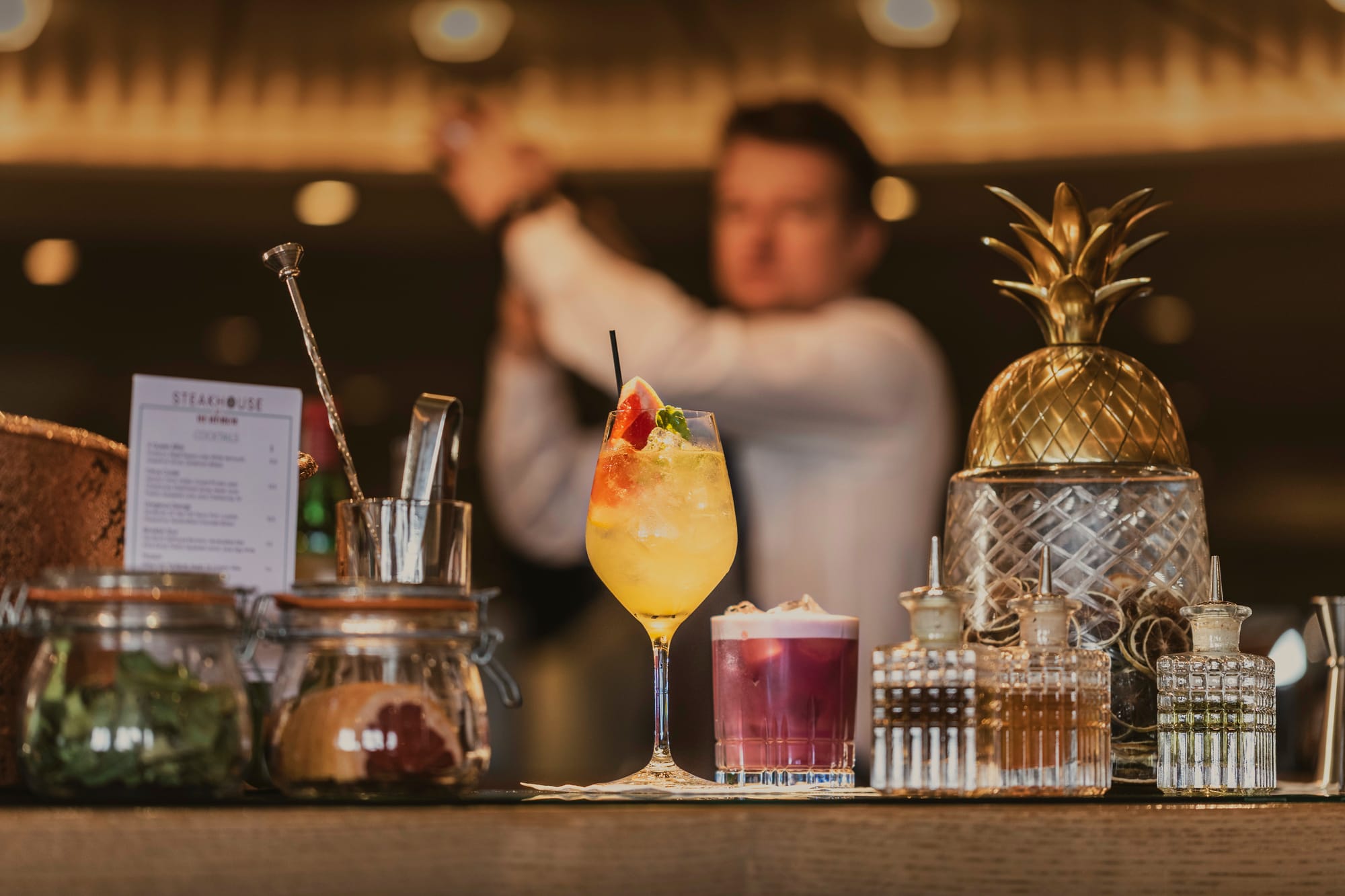 Several cocktails on a bar with a bartender mixing a drink in the background