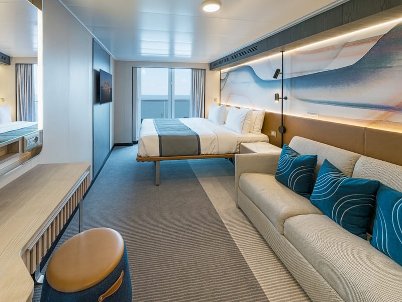 A bed in a cruise cabin with a sofa and a balcony